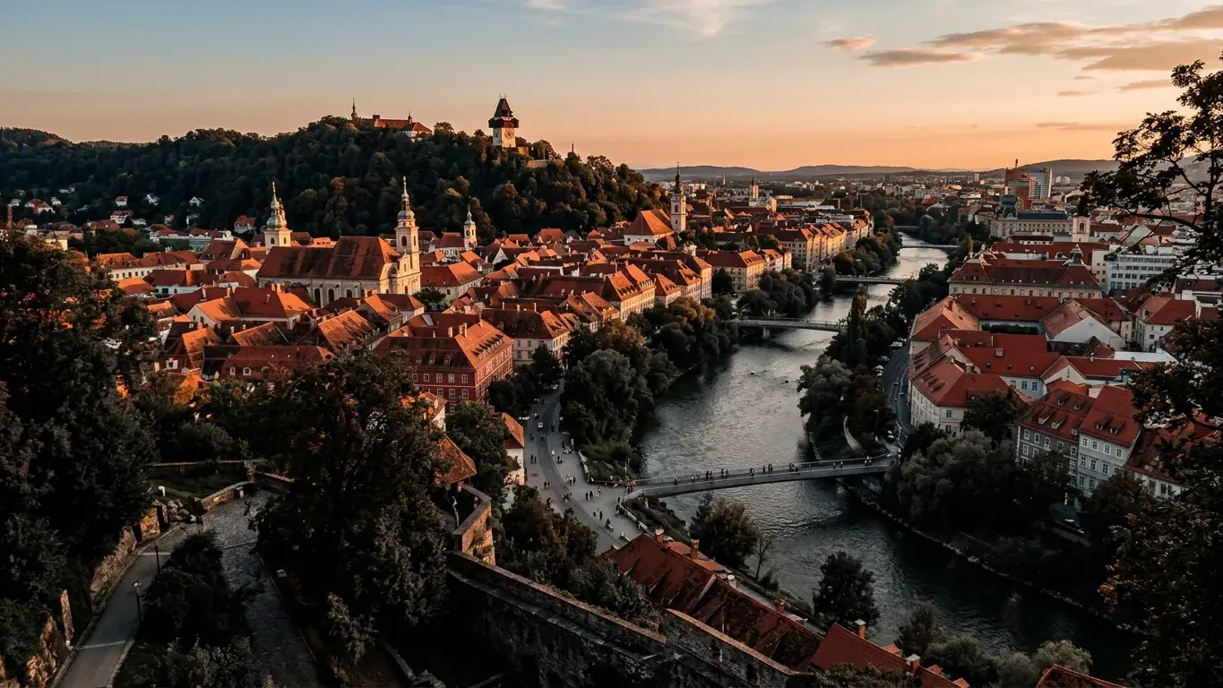 Blick über die Dächer der Grazer Altstadt mit Uhrturm auf dem Schlossberg und der Mur im Abendlicht